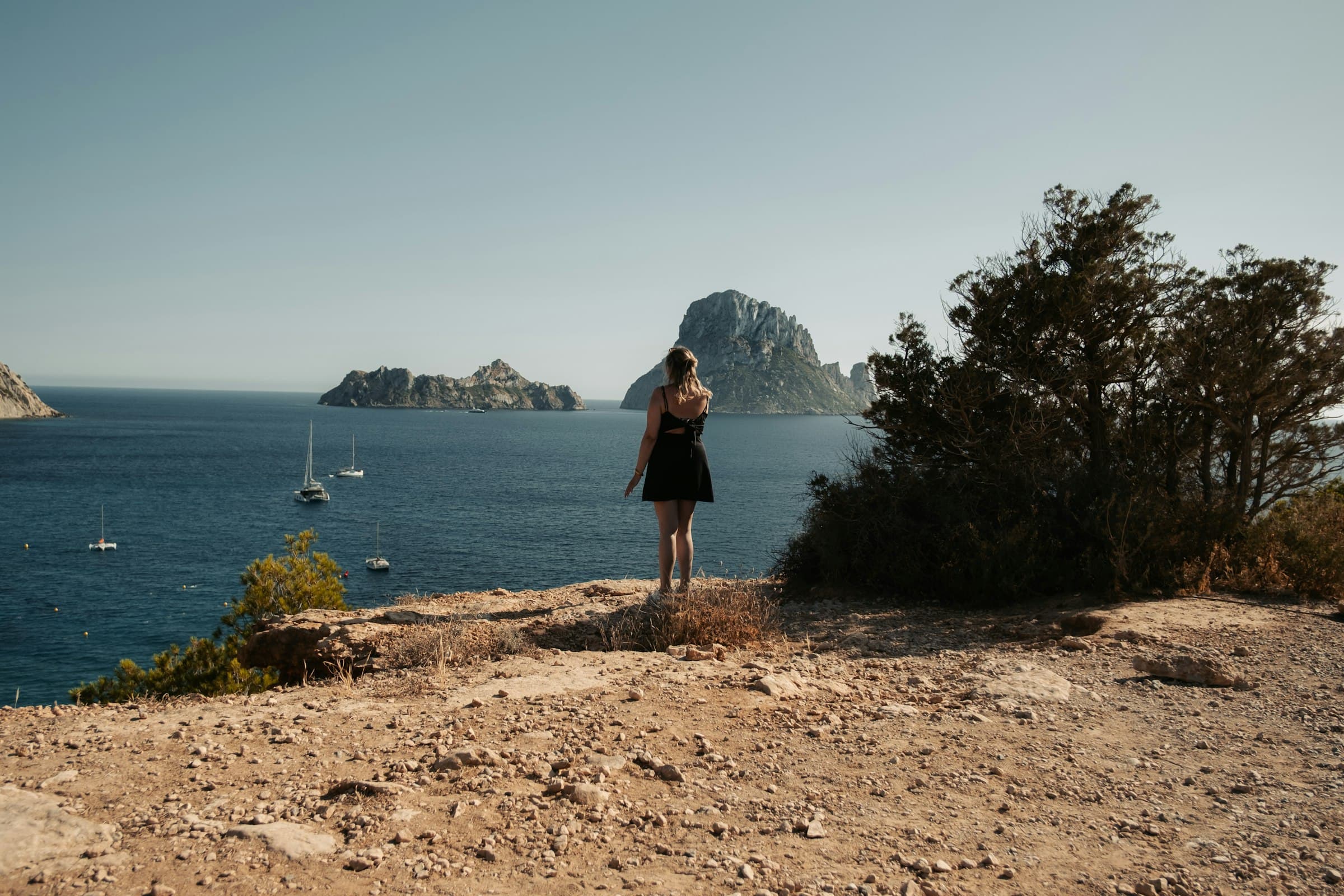 A woman standing at a cliff edge gazes out over distant islands and sailboats at sea