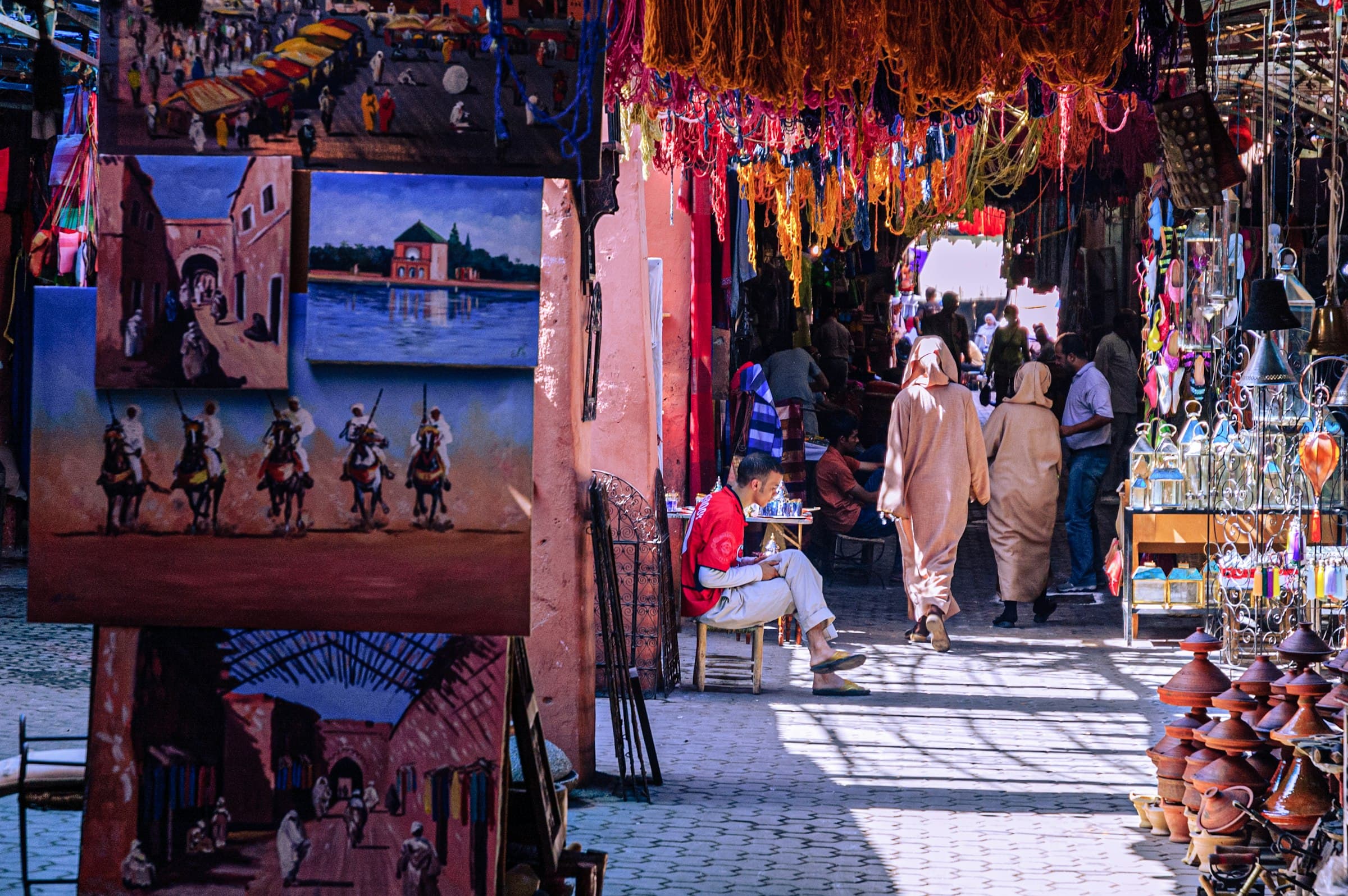 Shoppers in traditional djellabas walking through a sunlit souk alley in the Marrakech medina, lined with paintings, dyed wool and pottery