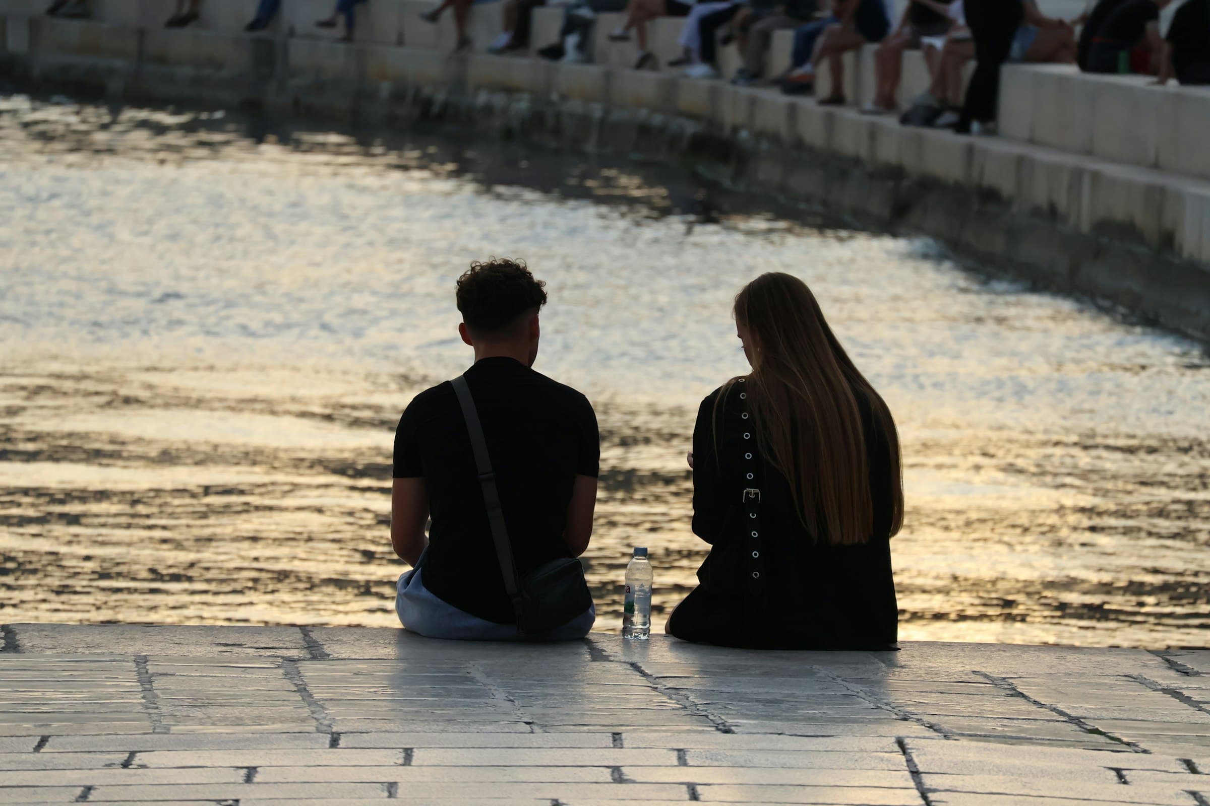 A couple sitting together with their backs to the water, watching a golden sunset