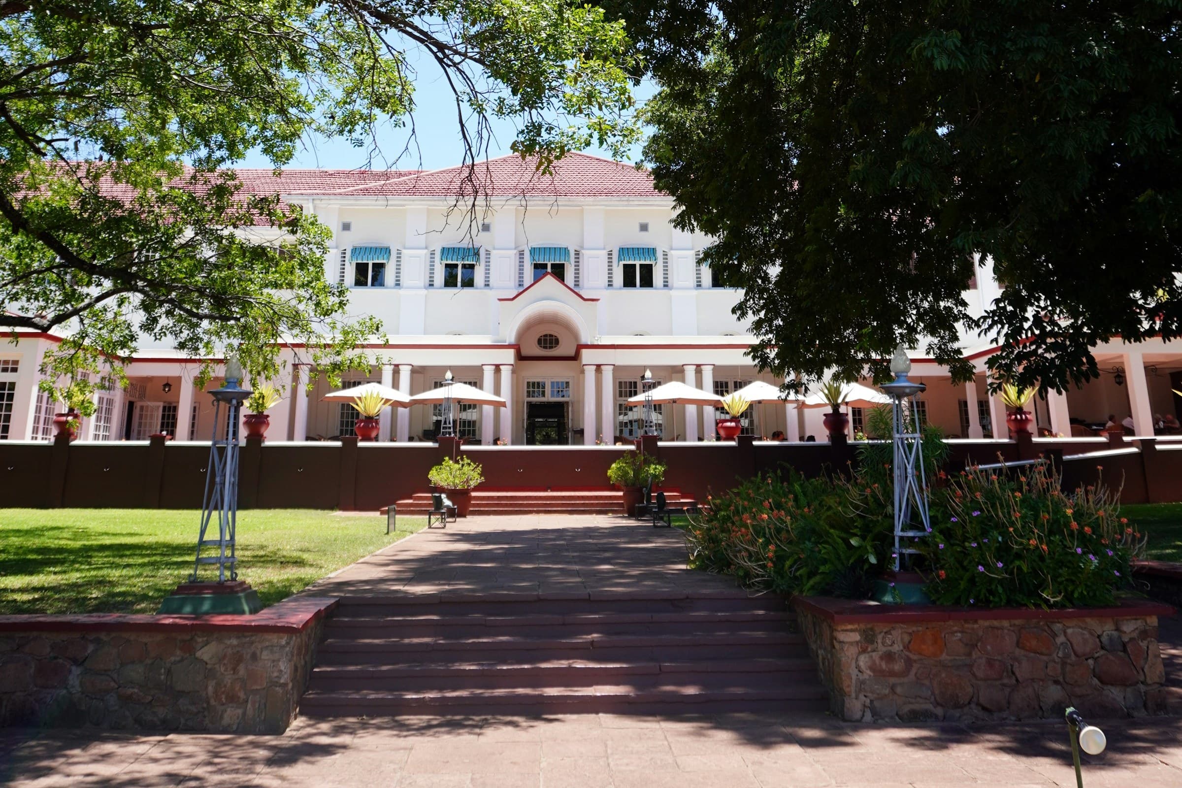 The white Edwardian facade and veranda of The Victoria Falls Hotel seen from its gardens, framed by large shade trees